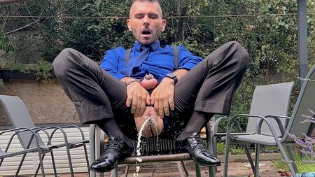 一位穿着西装、阴茎被锁住的男人正玩弄着自己的肛门，并穿着透明袜在公共场合小便